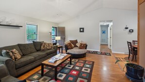 A spacious living room with a large sofa, a coffee table, and a patterned rug. There is a ceiling fan, a window with blinds, and a door leading to another room. - Whispering Pines - Spectacular Views & Hot Tub 3 Bedroom Home (Burnsville)