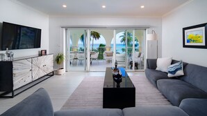 A modern living room with a flat-screen TV, grey sofas, a coffee table, and a view of the ocean through large glass doors.