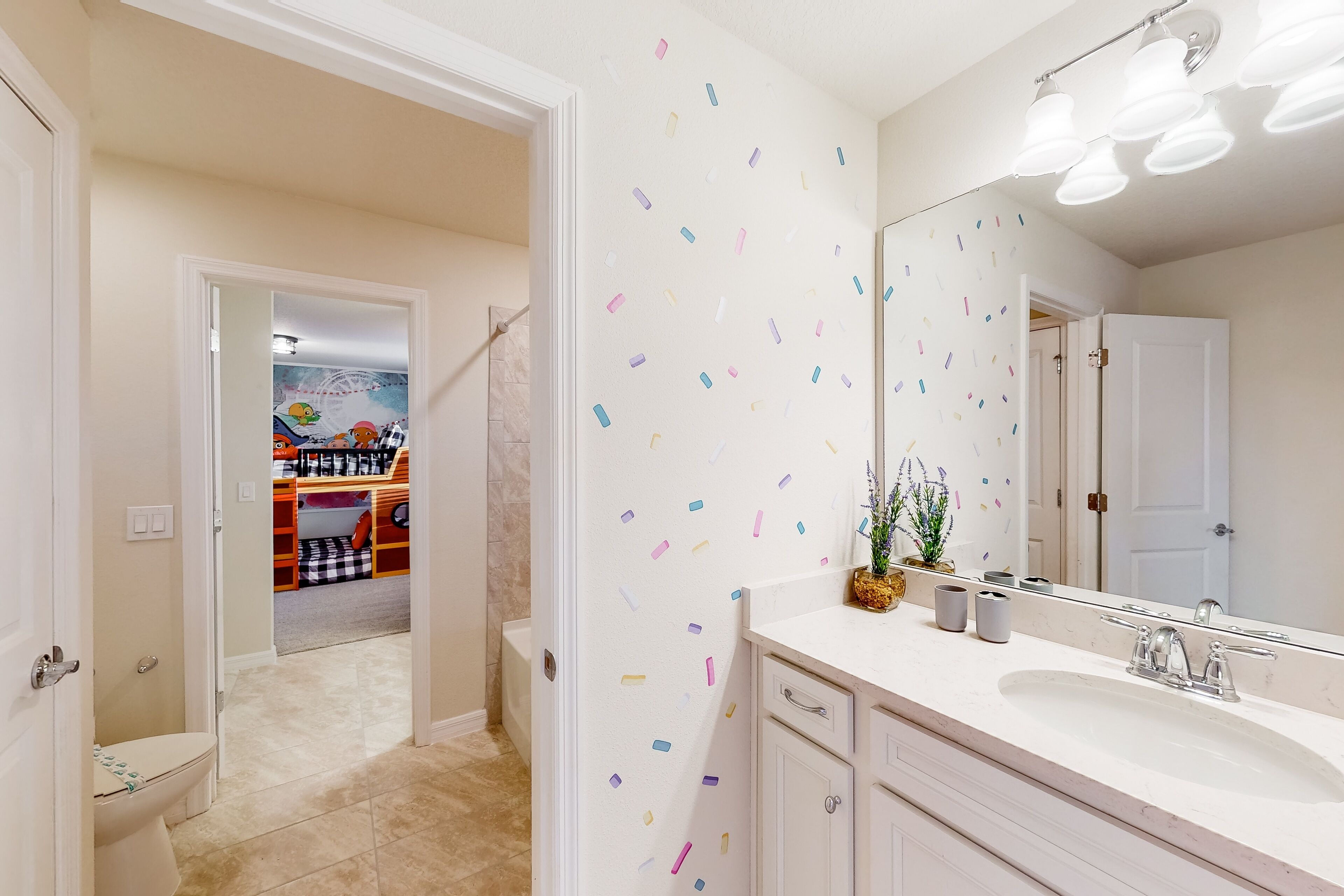 Une salle de bain avec un lavabo blanc, un miroir et des luminaires. On aperçoit une zone de douche à l’arrière-plan.