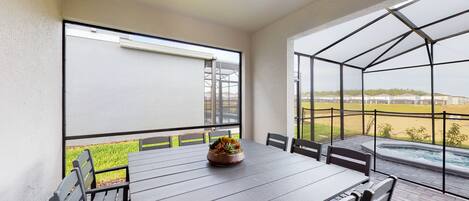 En overdækket terrasse med et bord og stole, en ventilator i loftet samt udsigt til en swimmingpool og grøn bevoksning.