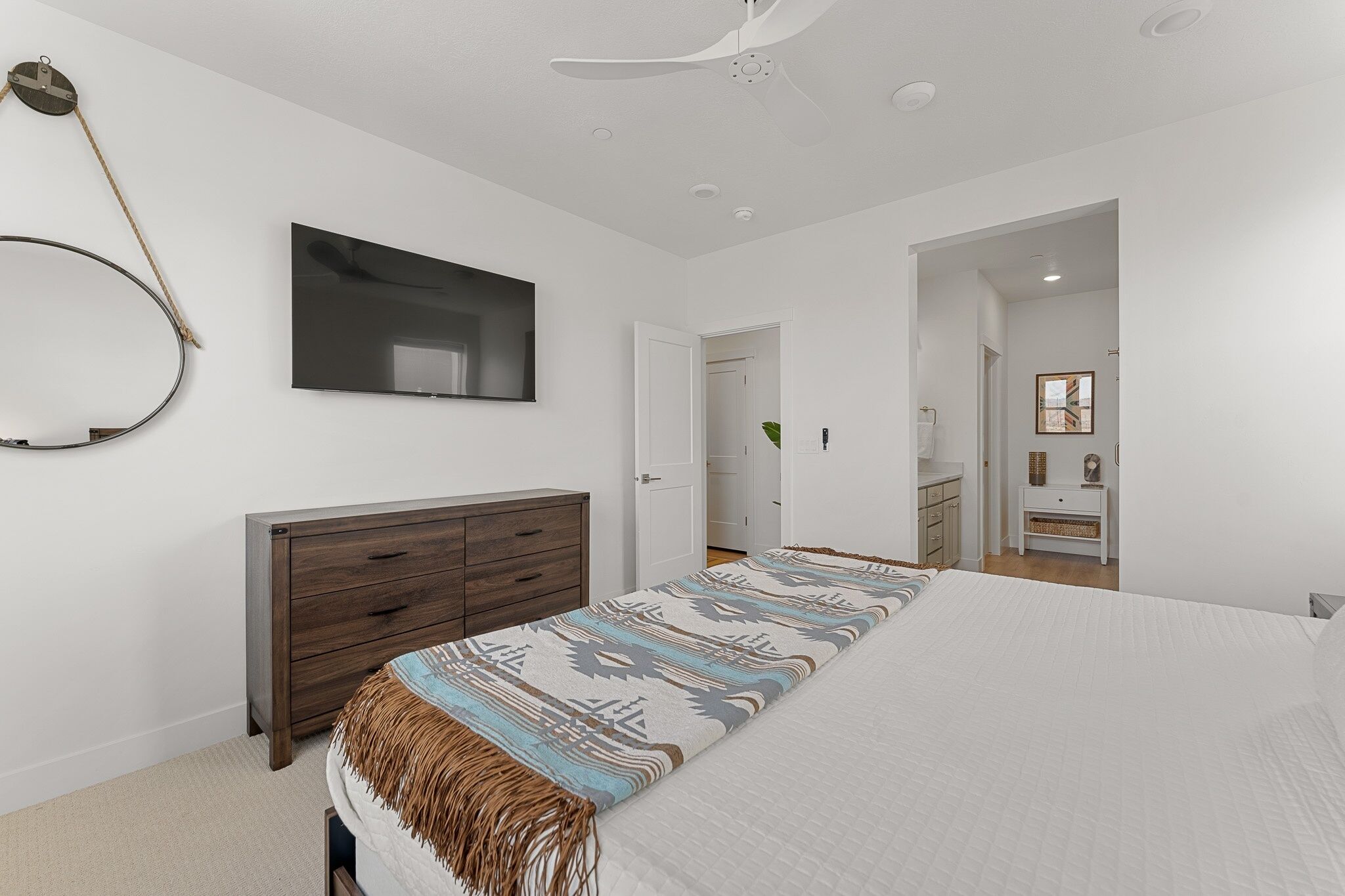 A bedroom with a bed, a wooden dresser, a ceiling fan, a wall-mounted TV, and a door leading to another room.