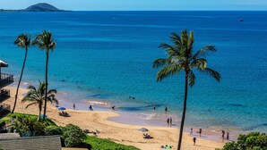 A beach with palm trees, clear blue water, and a distant mountain. - Maui Kamaole L107 1 Bedroom Condo (Kihei)