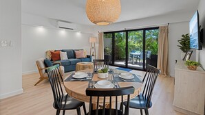 A modern living room with a dining area, large windows, and a TV mounted on the wall. - Maui Kamaole L107 1 Bedroom Condo (Kihei)