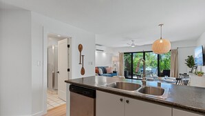 A modern kitchen with a dishwasher, double sink, and a dining area with a table and chairs. - Maui Kamaole L107 1 Bedroom Condo (Kihei)