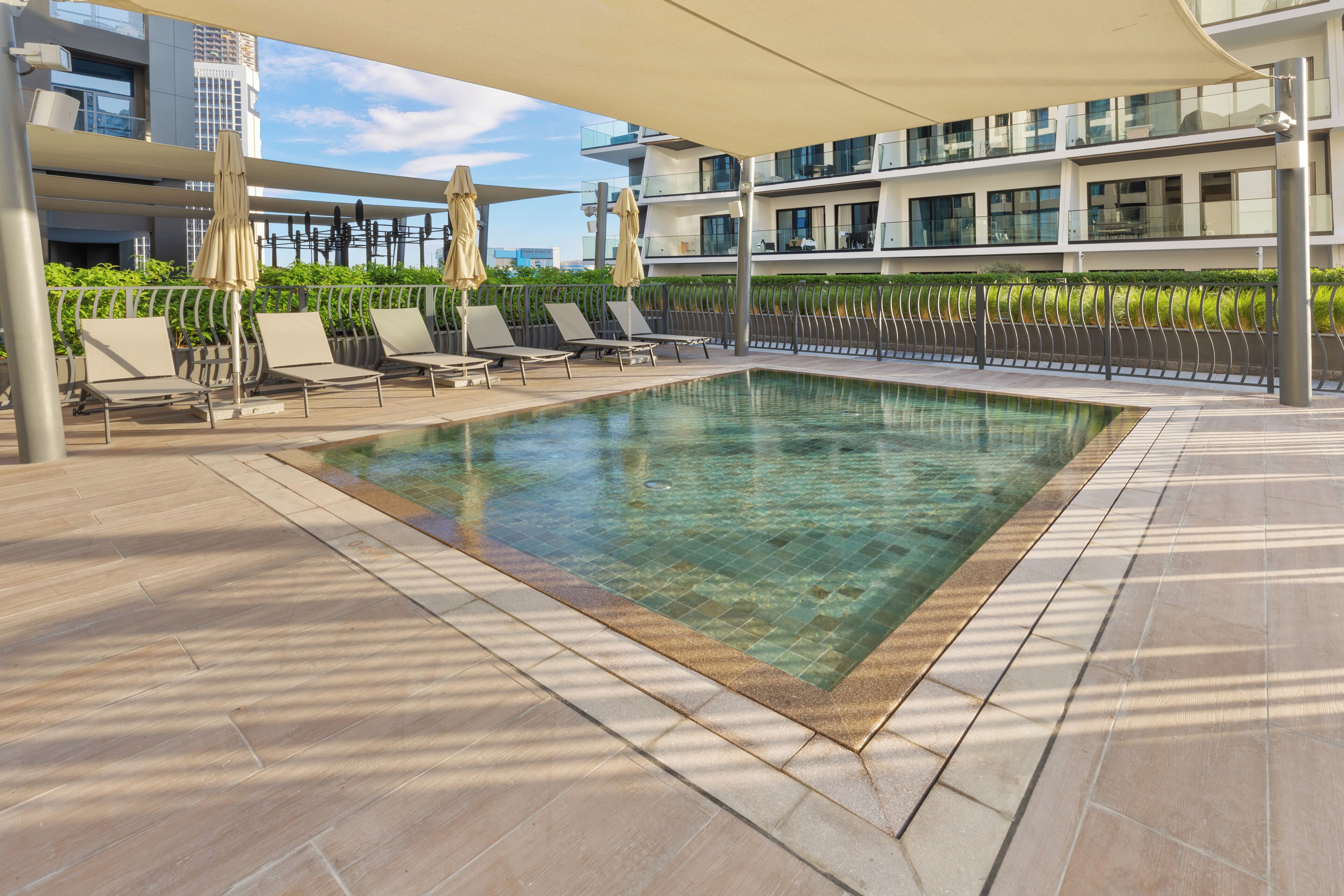 A modern outdoor swimming pool with lounge chairs and umbrellas, surrounded by a metal fence and a building with balconies.