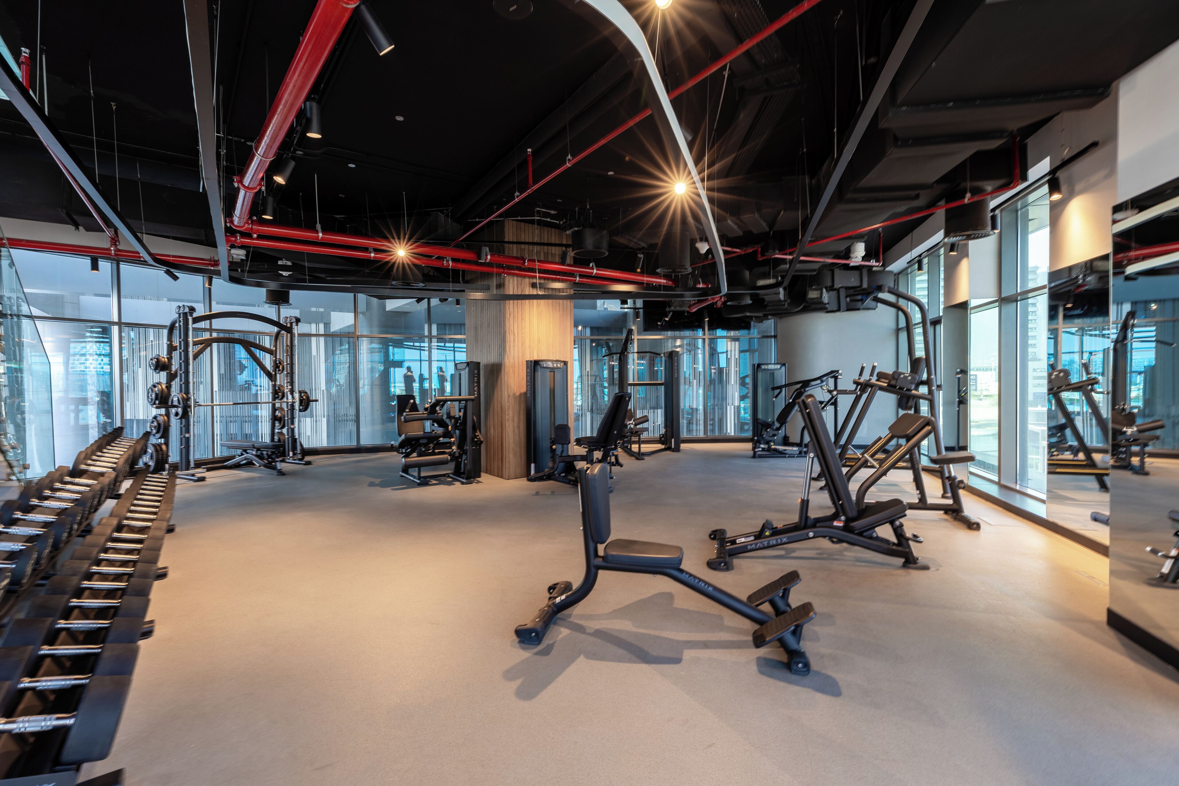 A modern gym with various exercise equipment, large windows, and a high ceiling with exposed pipes.