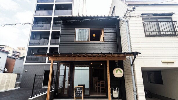 A two-story building with a wooden pergola entrance, a sign indicating a guesthouse, and a small window with a light fixture. - Fukuoka Guesthouse CAMP (Fukuoka)