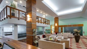 Um hall amplo com balcão de atendimento em madeira, áreas de estar confortáveis, escadaria e parede de vidro.