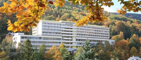 A multi-story building with a flat roof, surrounded by autumn foliage and a hilly backdrop.