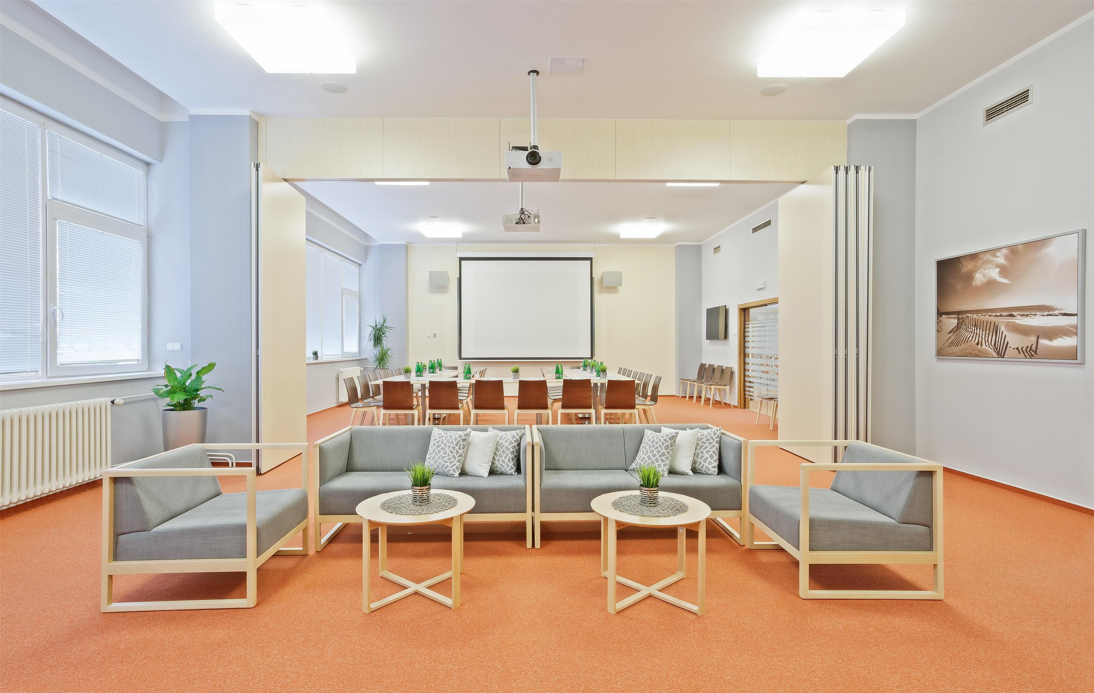 Un salón de conferencias con una pantalla grande, varias sillas, un sofá y pequeñas mesas con plantas.
