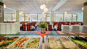 Uma cafeteria ampla com várias mesas e cadeiras, um buffet com diversas opções de comida e janelas grandes que oferecem vista para árvores.