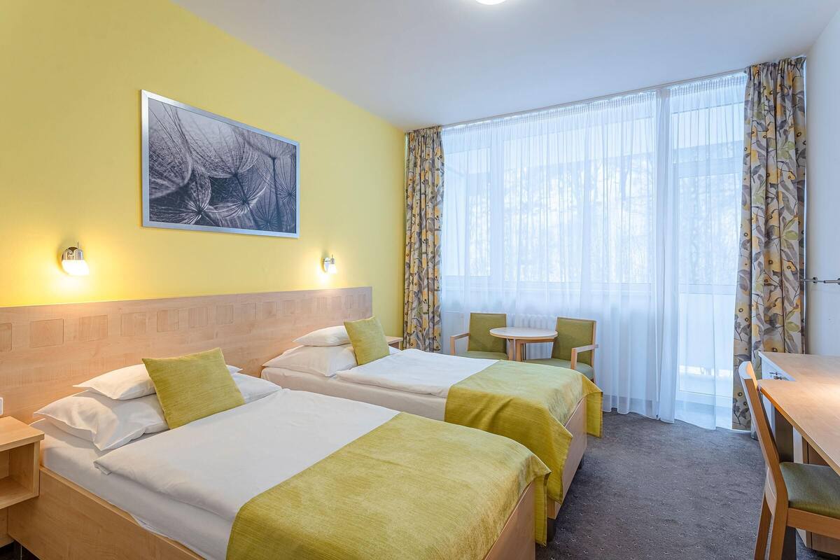 Ein Hotelzimmer mit zwei Betten, einem kleinen Tisch, einem Stuhl und einem Fenster mit Vorhängen.