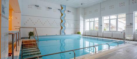 Una amplia piscina interior con una escalera recta que baja a la piscina, paredes de azulejos y grandes ventanales que permiten la entrada de luz natural.