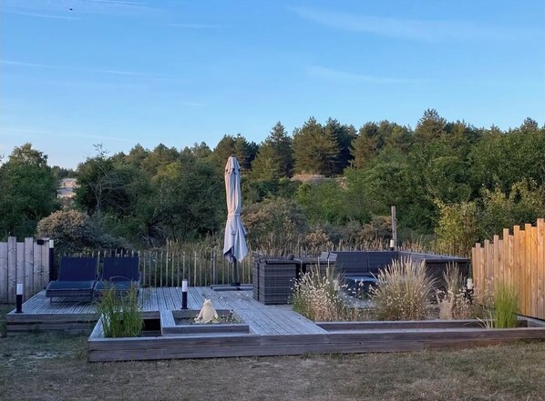 Una terraza con mobiliario y un área techada, rodeada por una valla de madera y follaje.