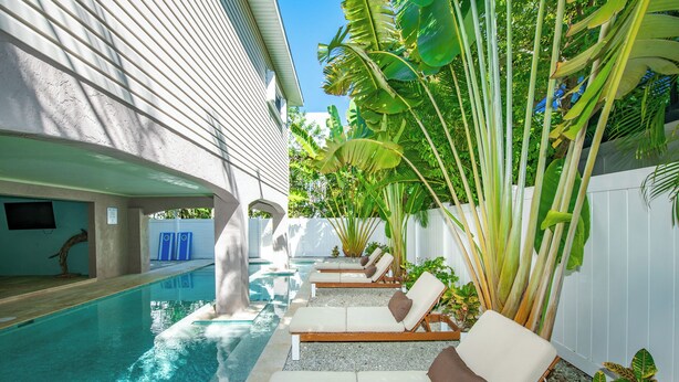 A poolside area with lounge chairs and a swimming pool, surrounded by tropical plants and a white fence.