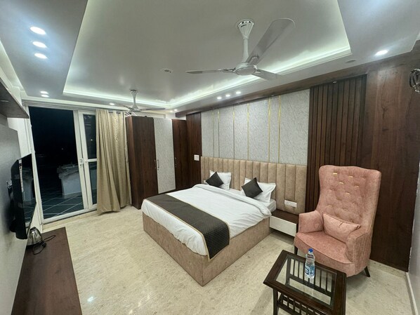 A modern hotel room with a large bed, a ceiling fan, and a seating area with a chair and table. - The Royal Oakes (New Delhi)