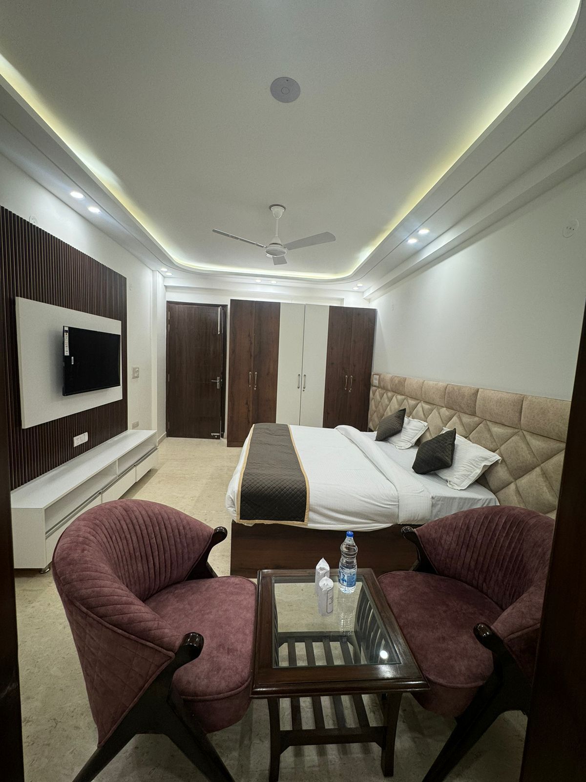 A modern hotel room with a bed, a TV, a ceiling fan, and a seating area with two chairs.