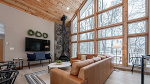 A modern living room with a large sofa, a flat-screen TV, a stone fireplace, and large windows offering a view of trees.