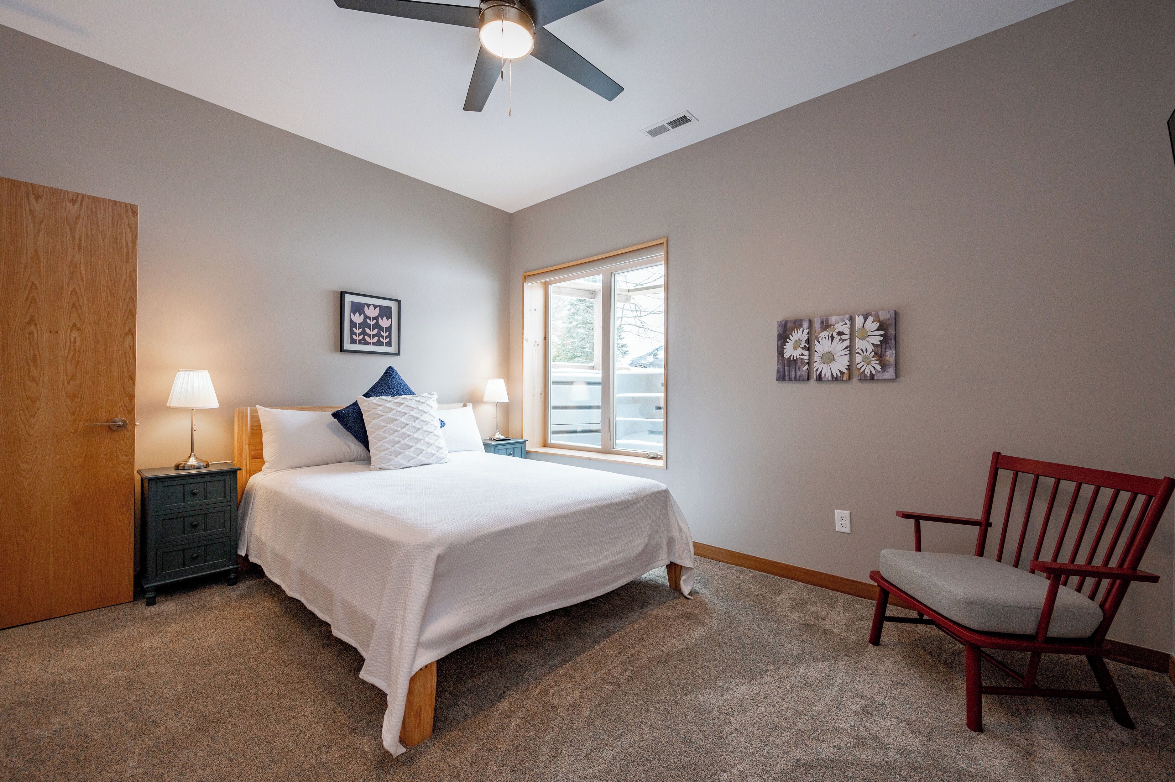A bedroom with a bed, nightstand, lamp, chair, and ceiling fan.