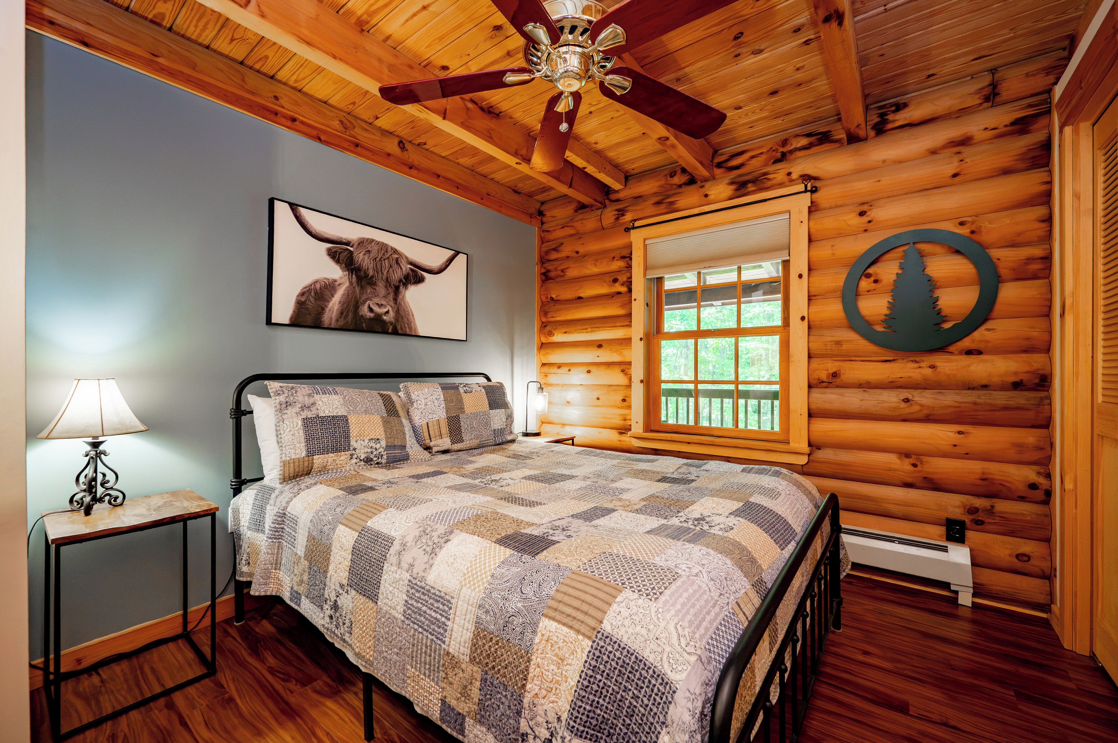 A log cabin bedroom with a bed, a ceiling fan, a window, a wall-mounted picture of a cow, and a circular wooden sign.