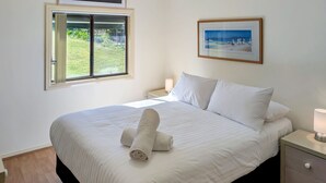 A neatly made bed with a rolled towel, a bedside lamp, and a framed picture on the wall. - Chill Out at the Basin by Experience Jervis Bay (Basin View)