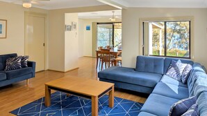 A modern living room with a blue sofa, a wooden coffee table, and a dining area with a table and chairs. - Chill Out at the Basin by Experience Jervis Bay (Basin View)