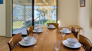 A dining area with a wooden table set for six, surrounded by chairs, with a view of greenery and trees through a glass door. - Chill Out at the Basin by Experience Jervis Bay (Basin View)