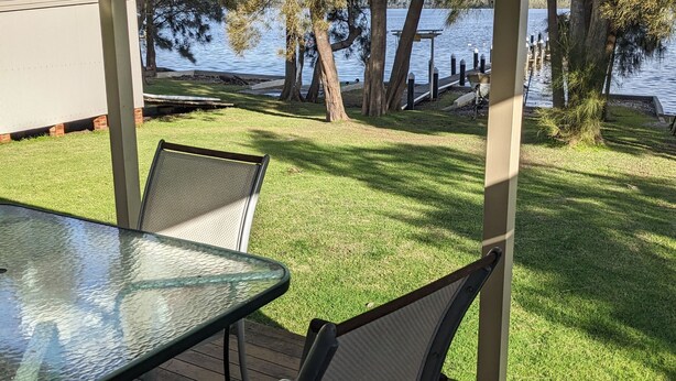 A covered patio with a glass table and chairs, overlooking a grassy area and a body of water. - Chill Out at the Basin by Experience Jervis Bay (Basin View)
