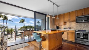 A modern kitchen with wooden cabinets, a large island, stainless steel appliances, and a view of palm trees.
