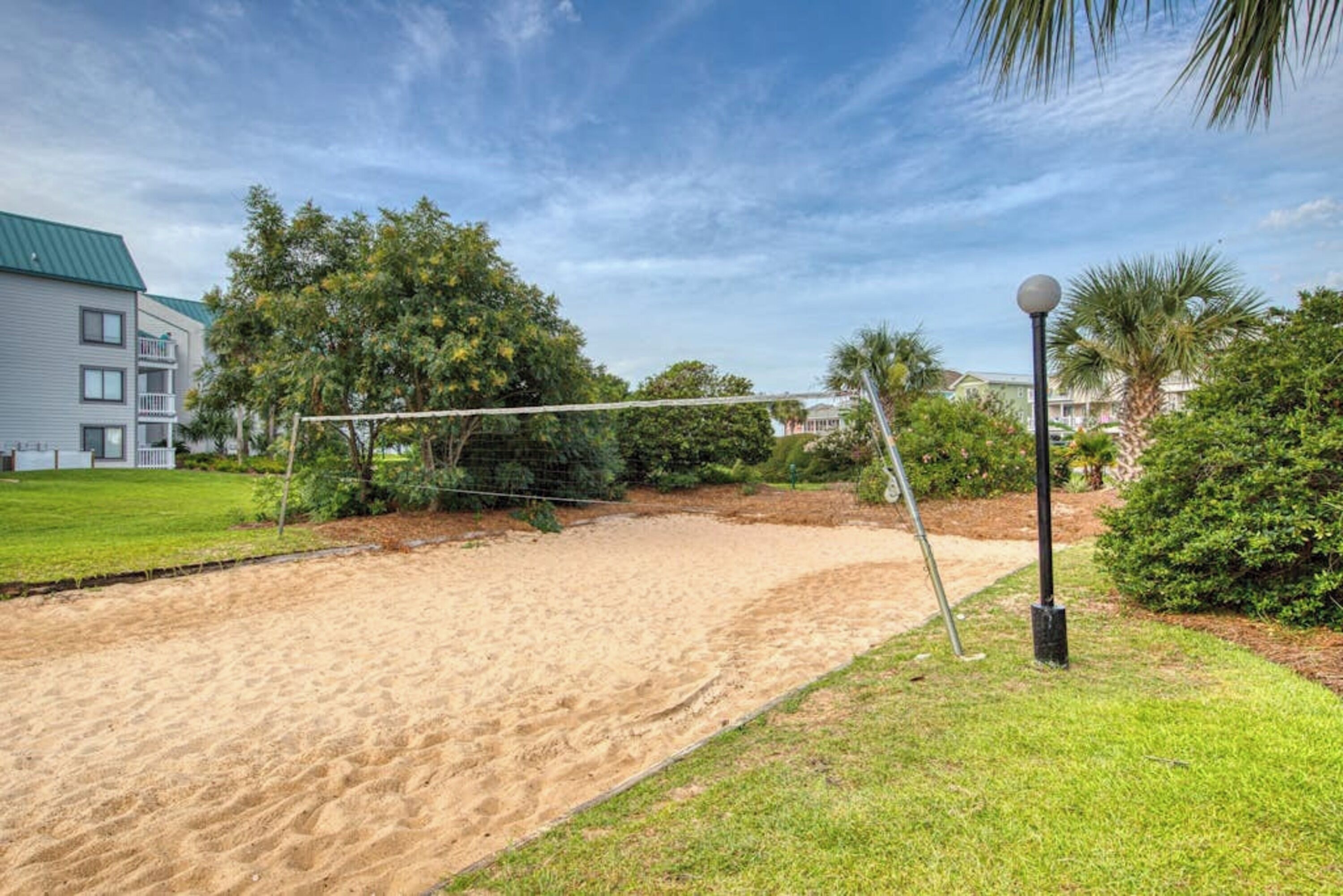 A sandy volleyball court with a net, surrounded by grass and trees, and a building with a green roof in the background.