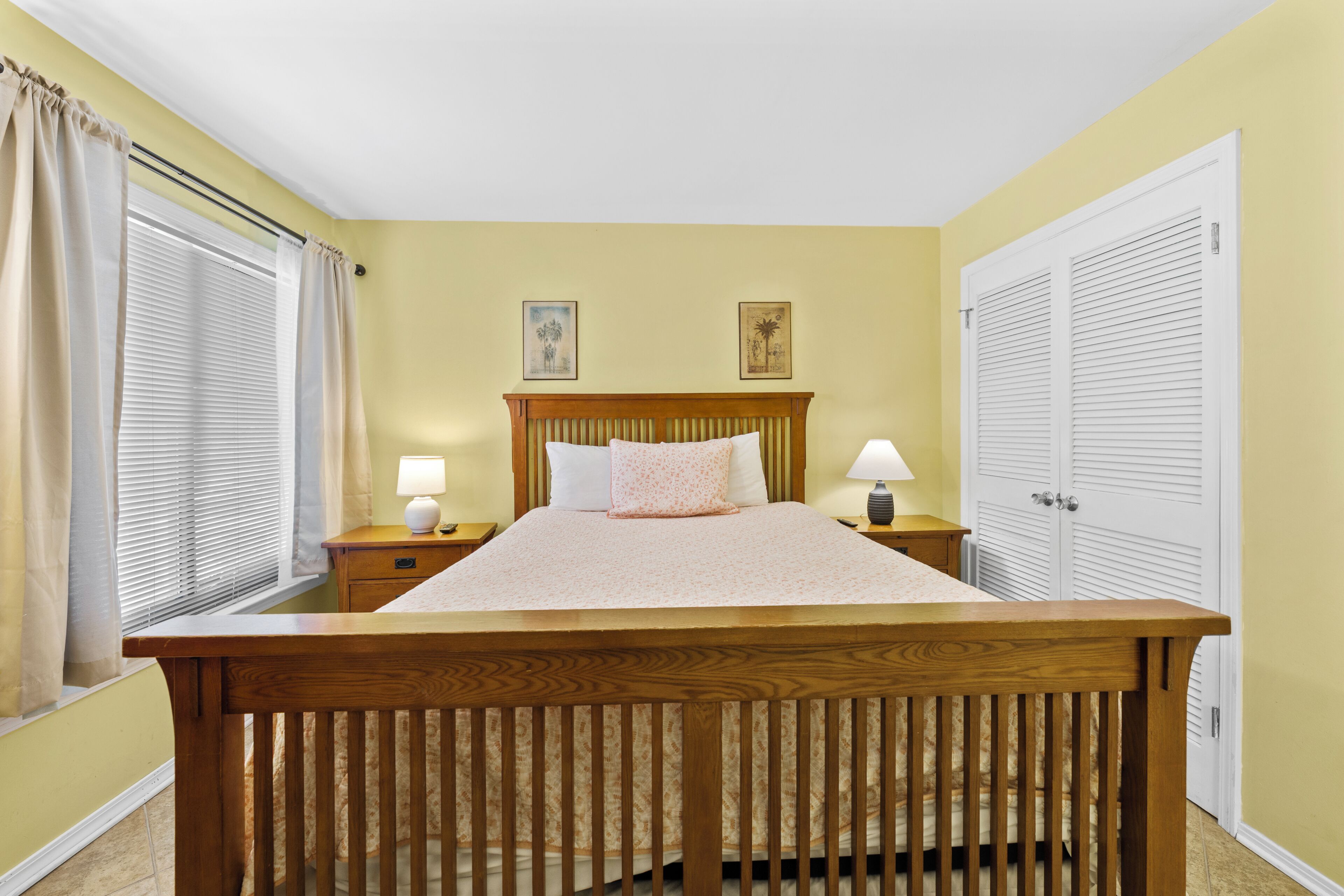 A bedroom with a wooden bed, two bedside tables with lamps, a window with blinds, and a door with louvered shutters.
