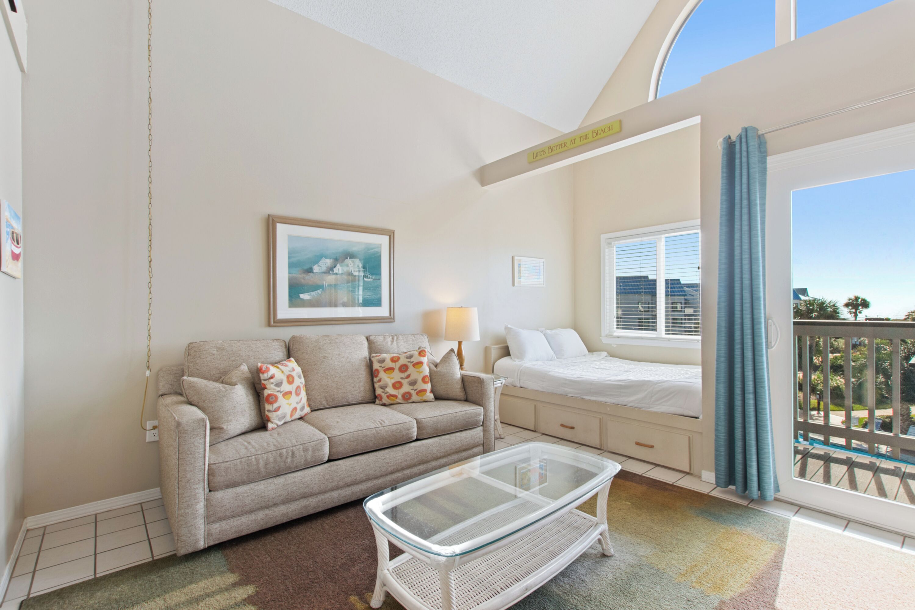A room with a sofa, a glass coffee table, a bed, a window with curtains, and a view of the outdoors.