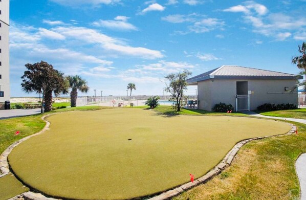 Un terrain de golf bien entretenu, doté d’un green d’entraînement, entouré d’une clôture, de palmiers et d’un petit bâtiment avec un toit en métal.