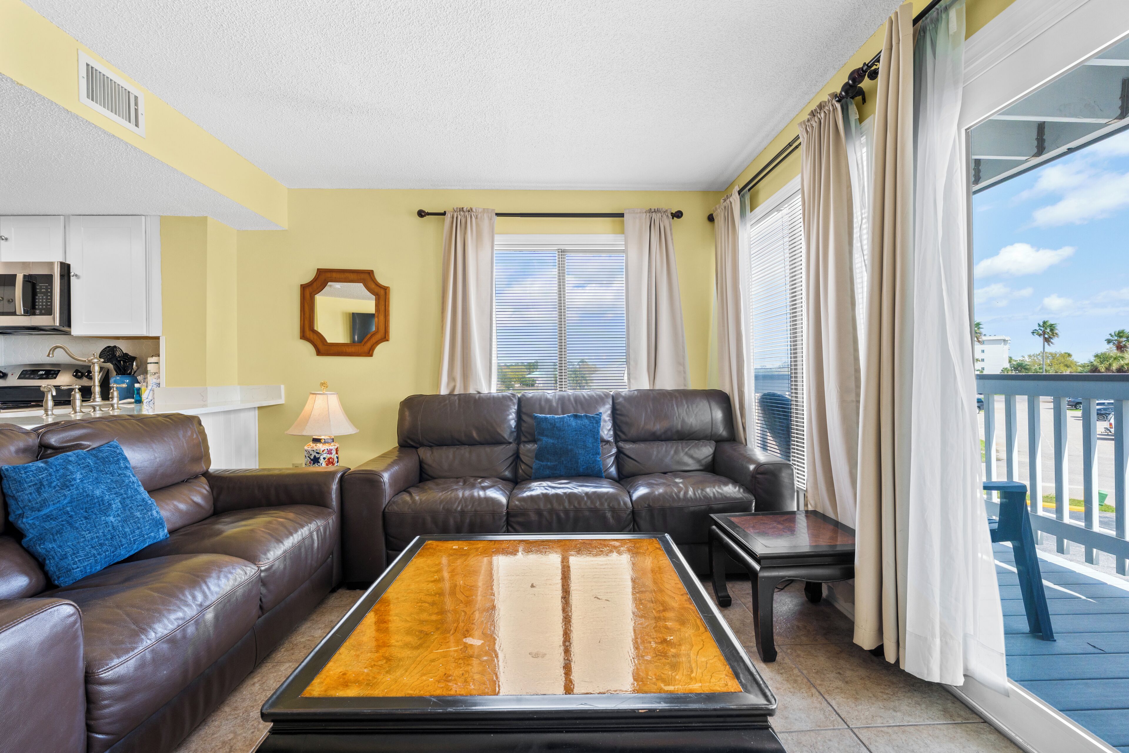 A living room with a brown leather sofa, a coffee table, a kitchen area with a microwave, and a balcony with a railing.