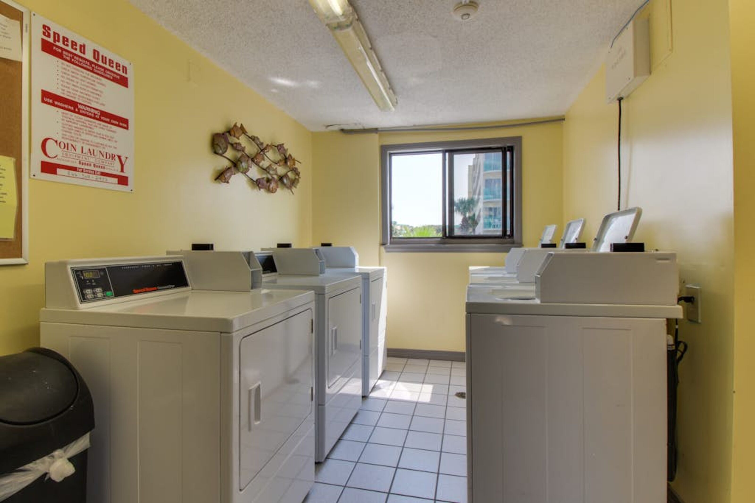 A laundry room with four washing machines, a coin laundry sign, and a window with a view of buildings.