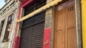 A building with a closed shutter door, a visible number '132' on the door, and a small window with a metal grid. - Hospedagem Rio Lapa  (Rio de Janeiro)