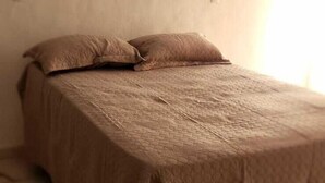 A bed with a textured bedspread and pillows, a plain wall, and a light source from the right. - Hospedagem Rio Lapa  (Rio de Janeiro)