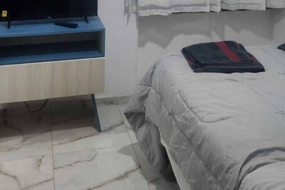 Un dormitorio con cama, televisor sobre un mueble, ventilador de techo y ventana con cortinas.