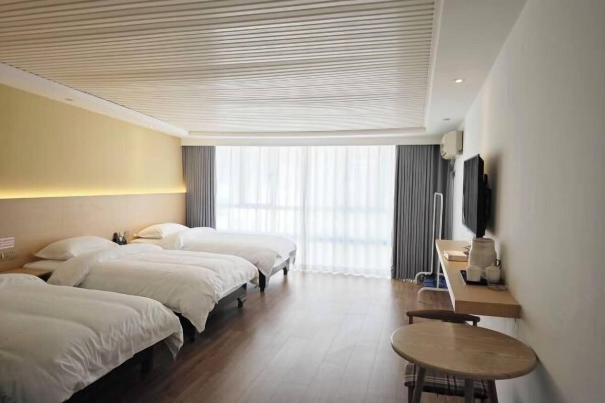 Habitación de hotel con tres camas, un escritorio y una silla, televisión y un amplio ventanal con cortinas.