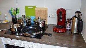 Una cocina con estufa, ollas, sartenes, cafetera y tabla de cortar.