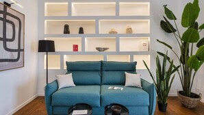 Un canapé de couleur turquoise avec deux coussins, un lampadaire noir et une grande plante en pot. Des étagères ornées de divers objets décoratifs se trouvent derrière le canapé.