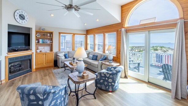 A spacious living room with a fireplace, a television, and a balcony with a view. - Magnificent oceanfront home in South Nags Head! (South Nags Head)
