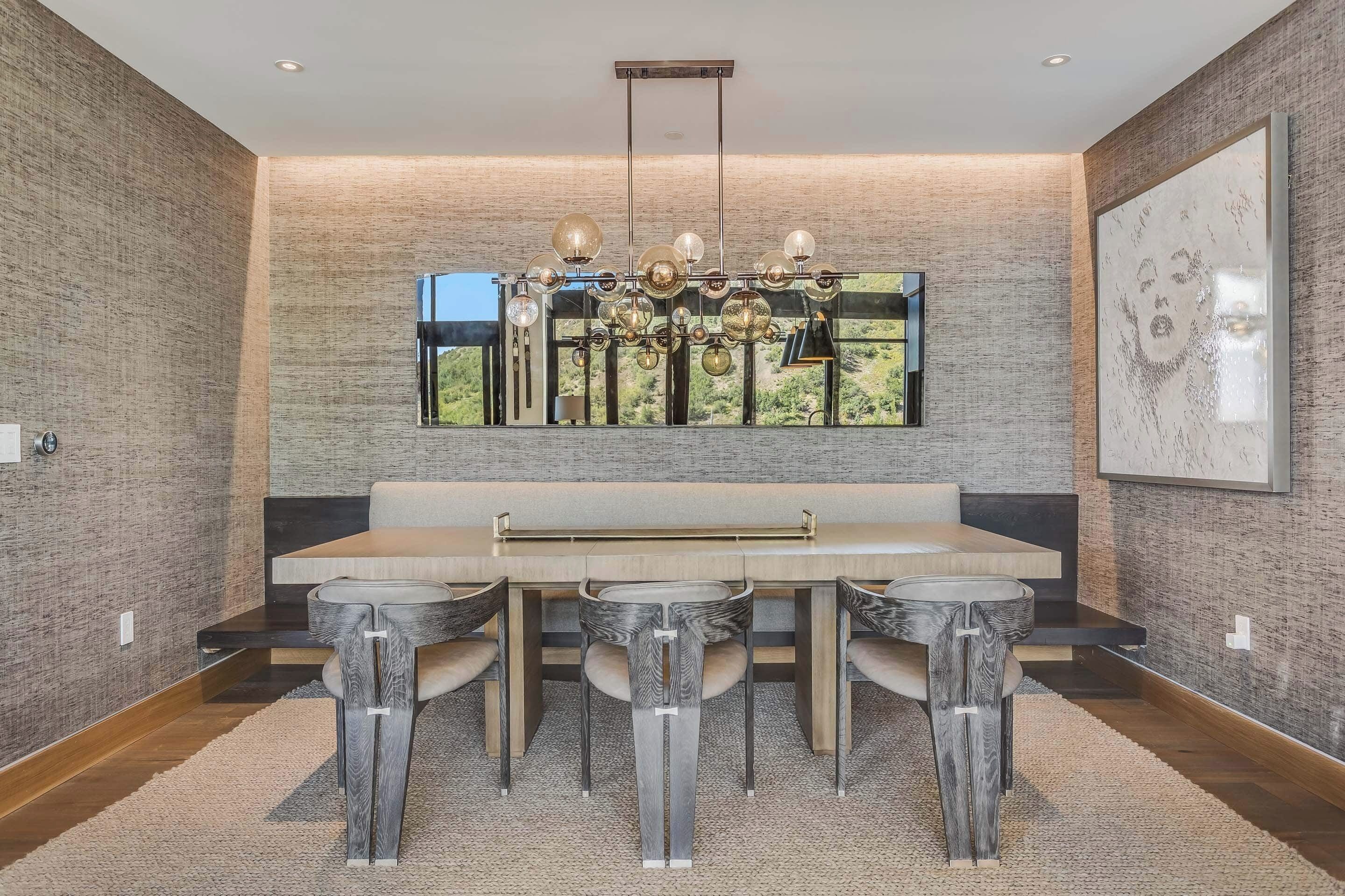 A modern dining area with a long table and chairs, a chandelier, and a large window with a view.