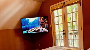 A bedroom with a bed, a television mounted on the wall, and a window with a view of trees. - Cozy Winston-Salem Vacation Cabin (Winston-Salem)