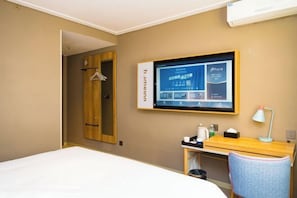 A hotel room with a bed, a desk with a lamp, a TV mounted on the wall, and a wardrobe.