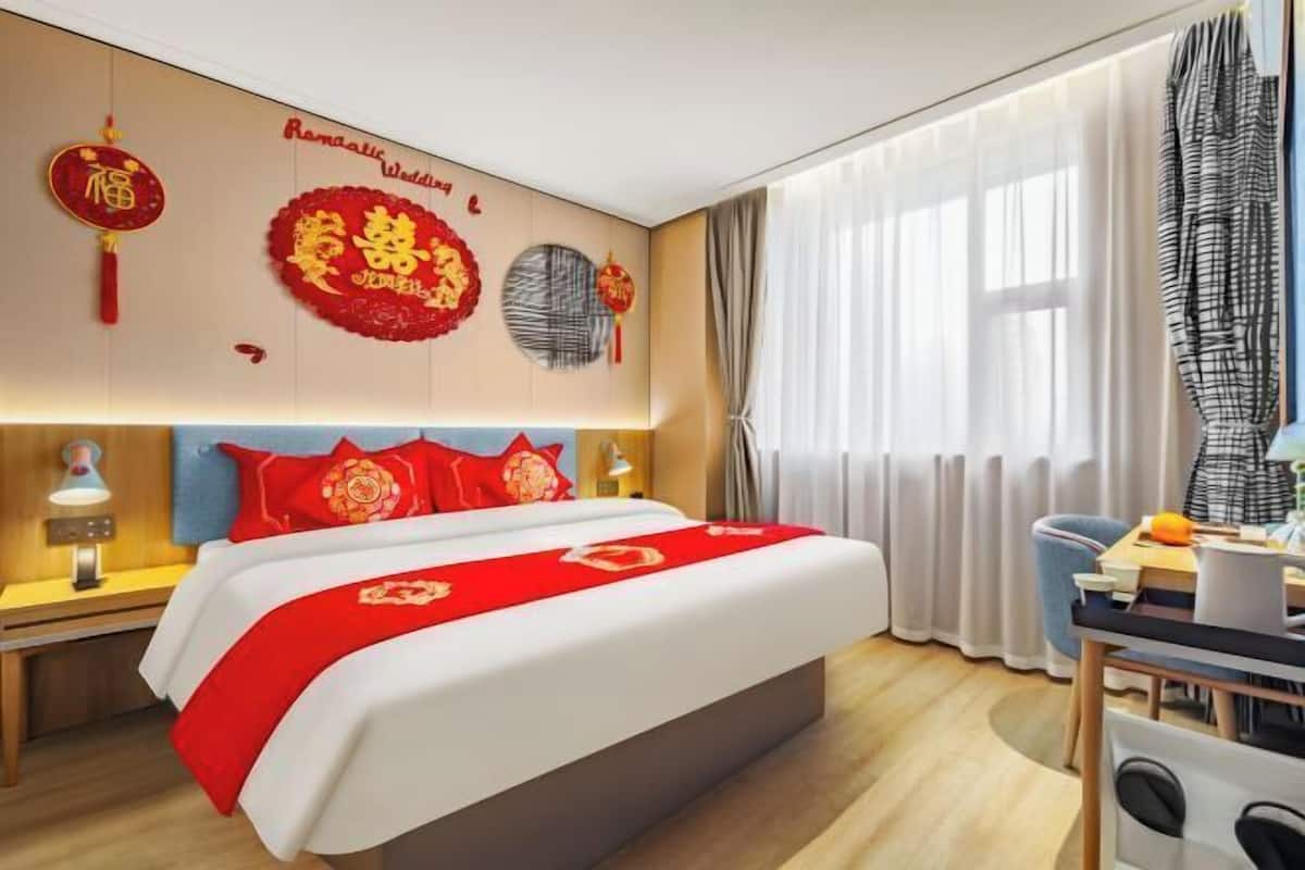 A hotel room with a bed, bedside tables, a desk with a chair, a window with curtains, and wall decorations.