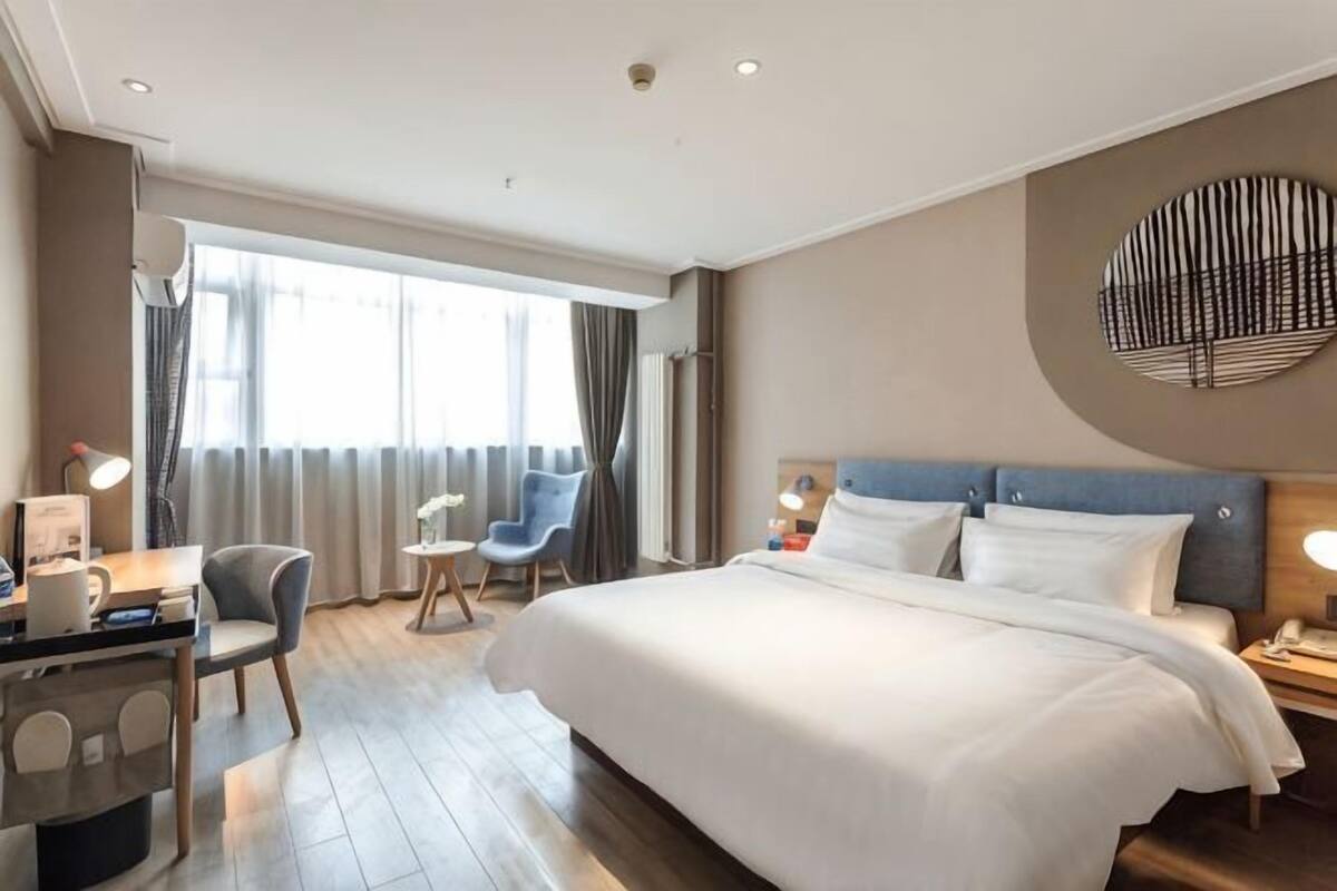 A hotel room with a large bed, a desk with a chair, a small table, and a chair. There is a window with curtains, a wall-mounted lamp, and a round wall decoration.