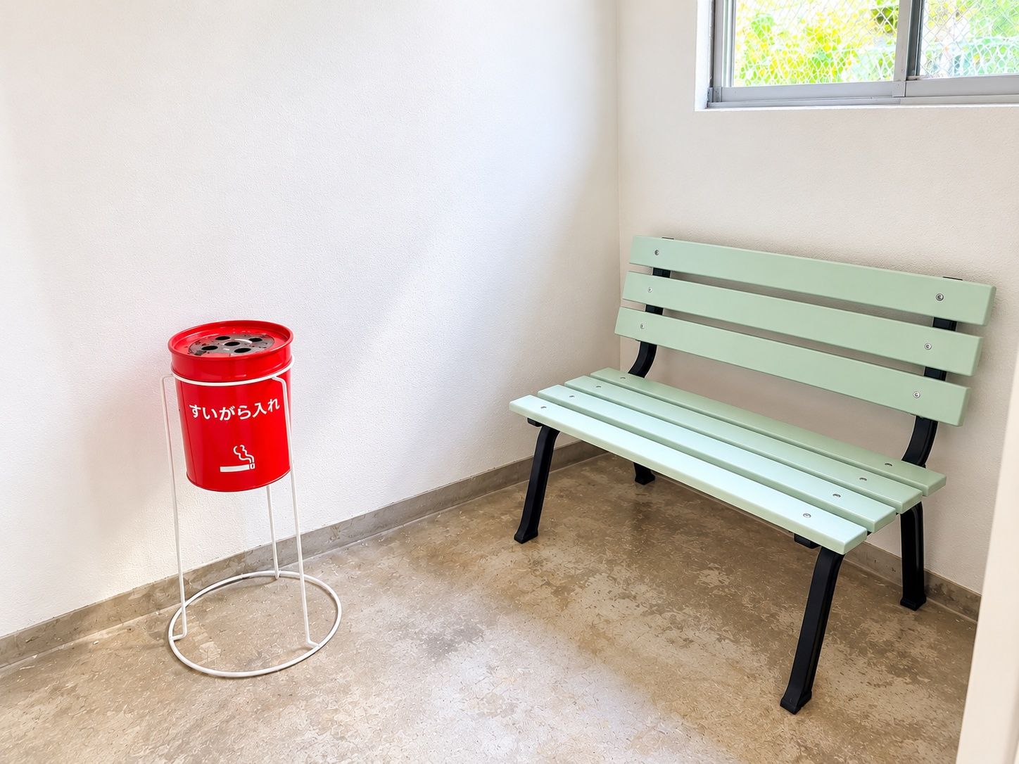 Une poubelle rouge avec des inscriptions en japonais, un banc vert et une fenêtre donnant sur un paysage verdoyant.