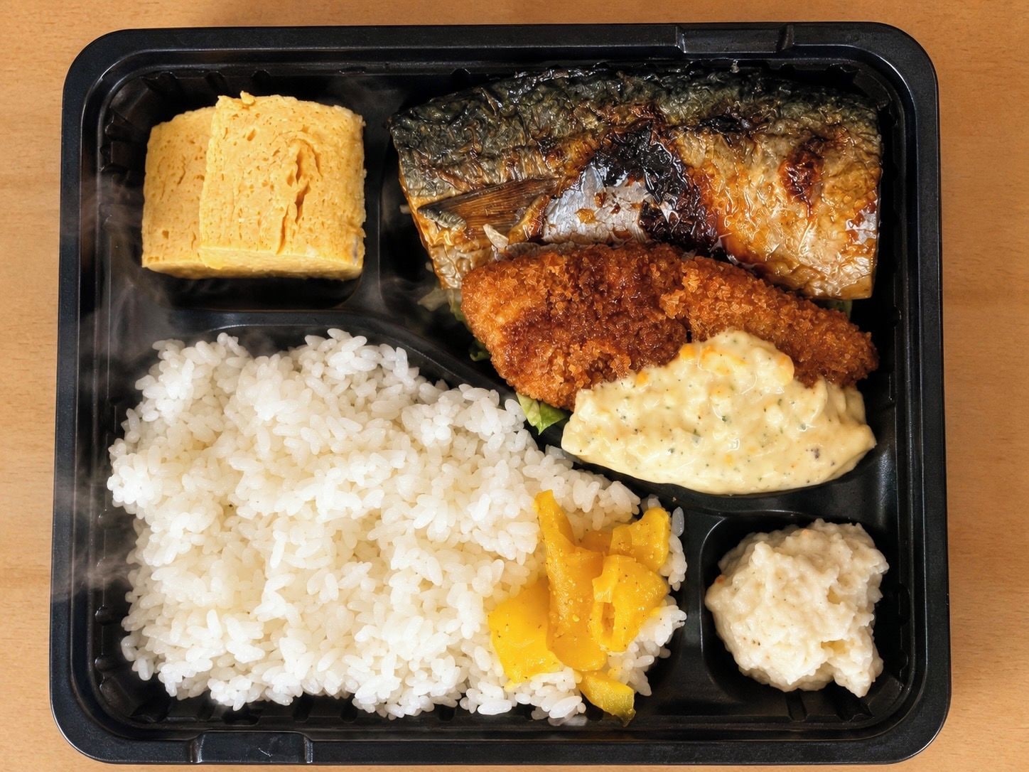 Una comida en un recipiente negro con arroz, un pescado, un elemento empanizado y salsa cremosa.