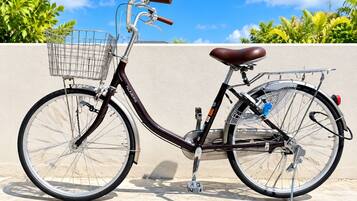 A stationary bicycle with a basket, a seat, and a rear rack, parked against a wall with a clear sky and greenery in the background.